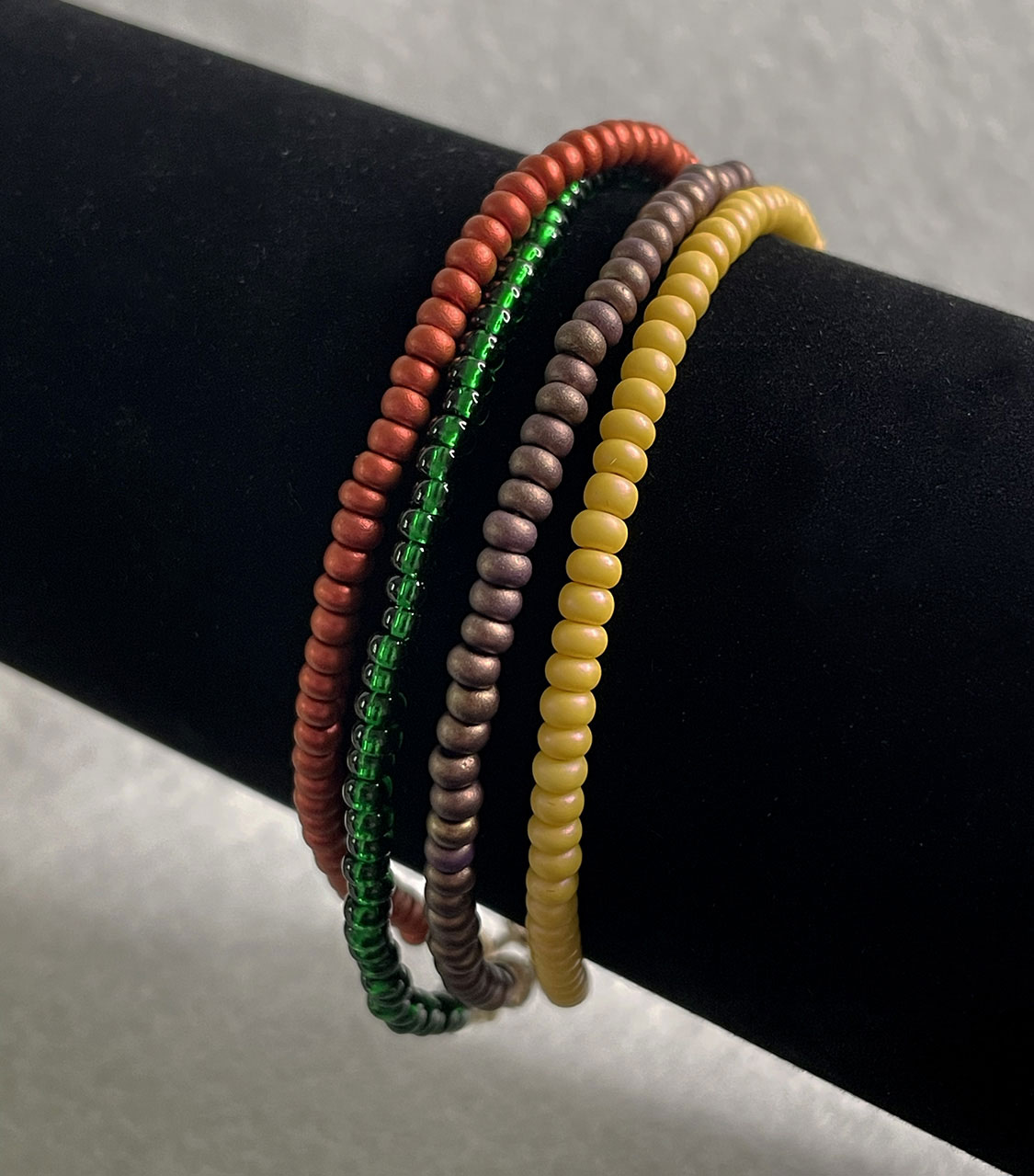 Four colorful beaded coil bracelets in red, green, brown, and mustard yellow wrapped around a black display stand.
